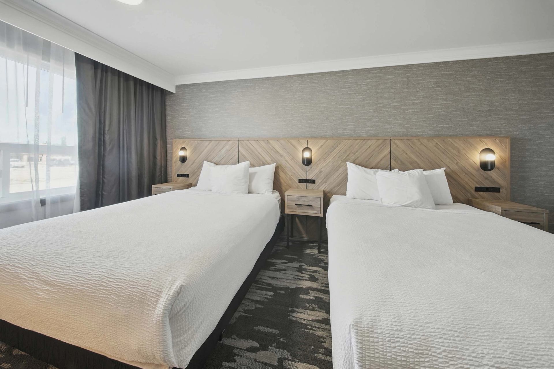 Hotel room with two double beds, wood headboard, bedside tables and wall lamps.
