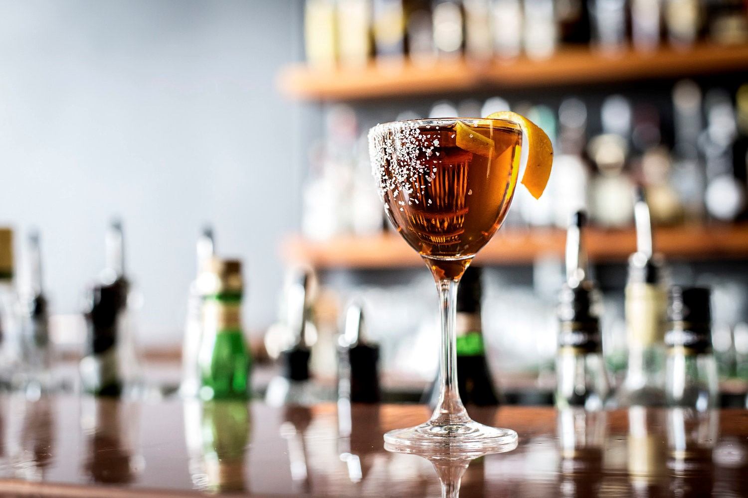 Amber cocktail with orange garnish on wood bar, bottles in background.