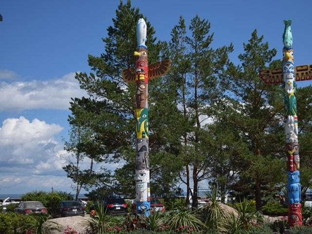 Colourful totem poles near trees and parked cars, with lake and blue sky.