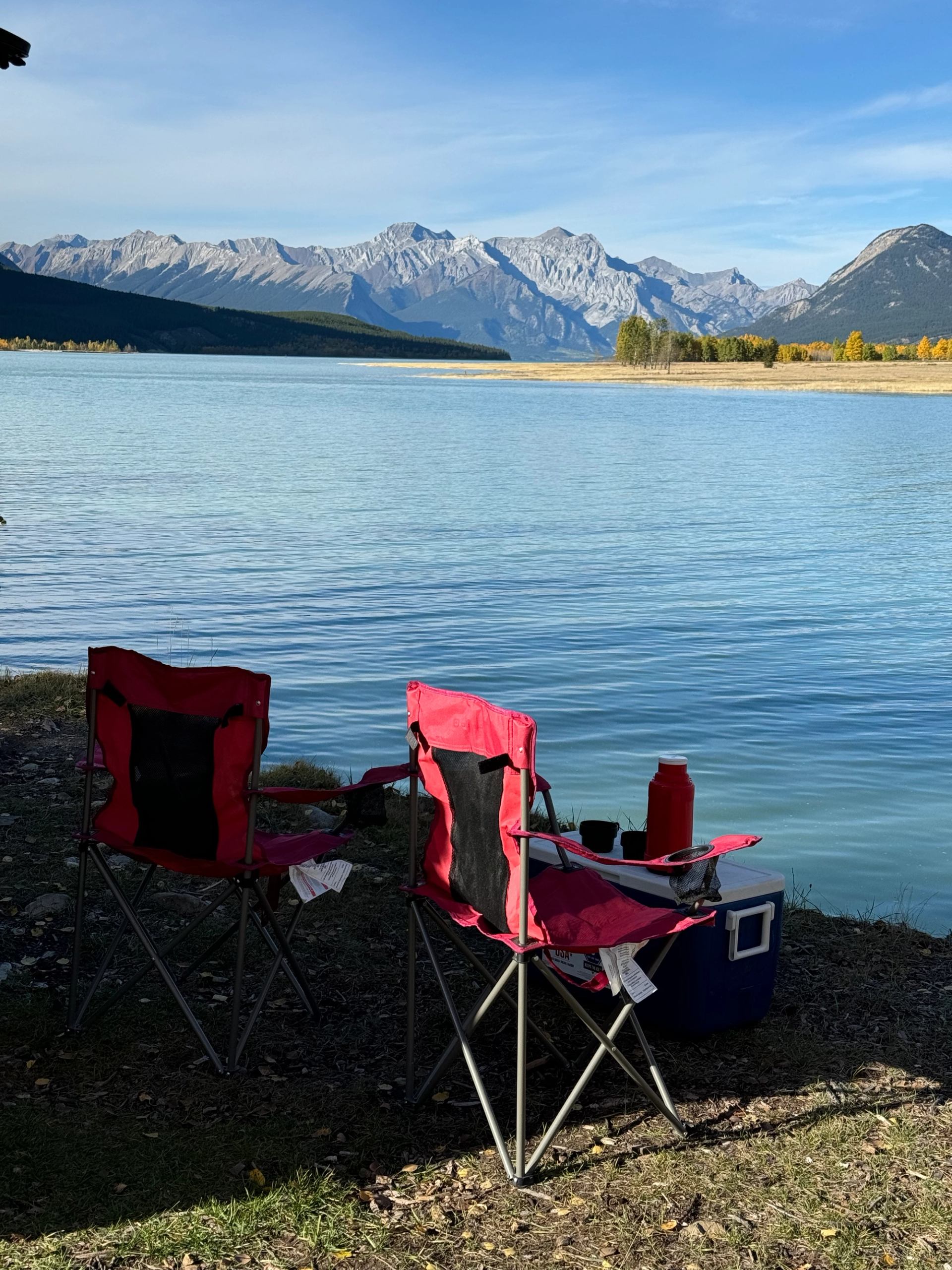 Two red camping chairs by a calm lake, facing distant rocky mountain range.
