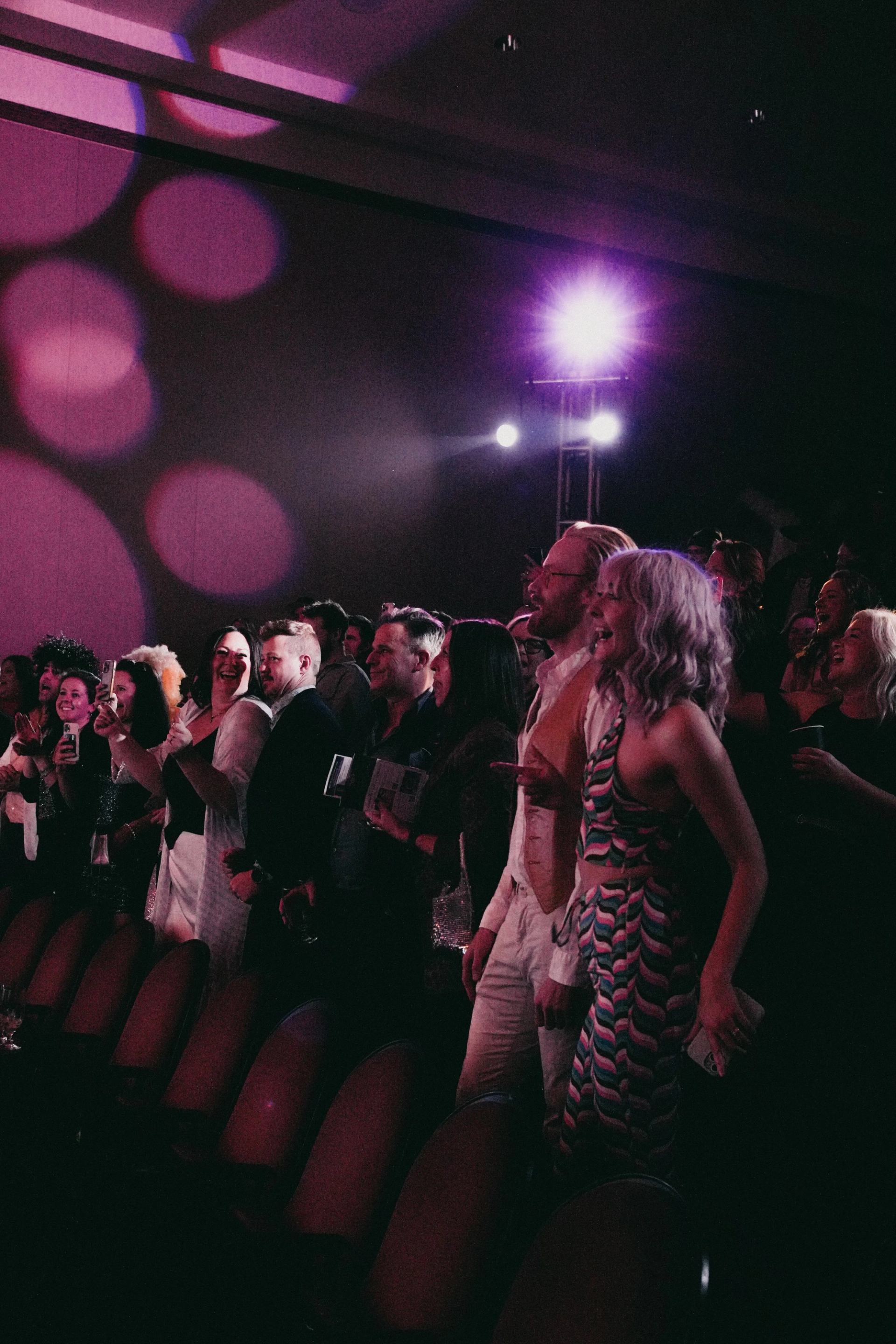 Audience members stand near theatre seats under purple stage lights, watching an unseen performance.