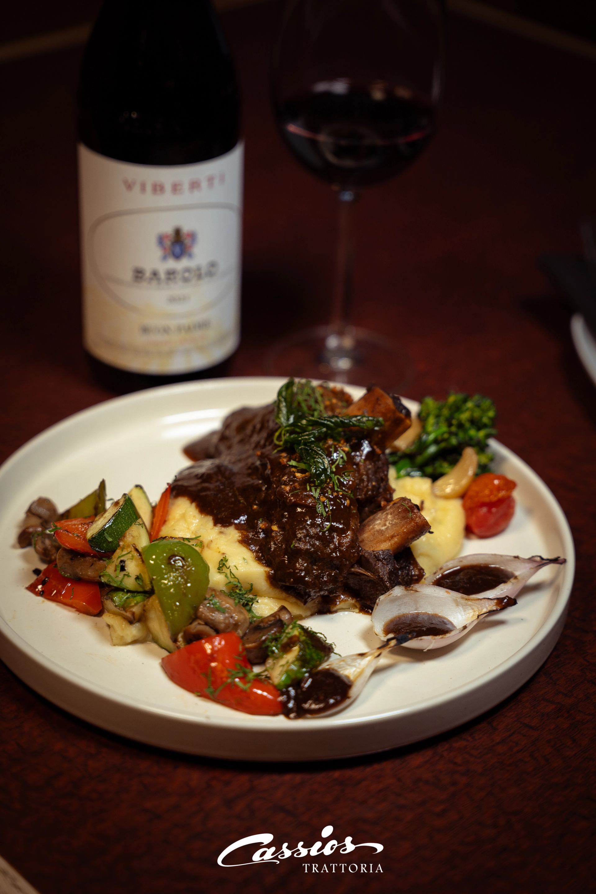 Braised meat on mashed potatoes with roasted vegetables, red wine and Barolo bottle.