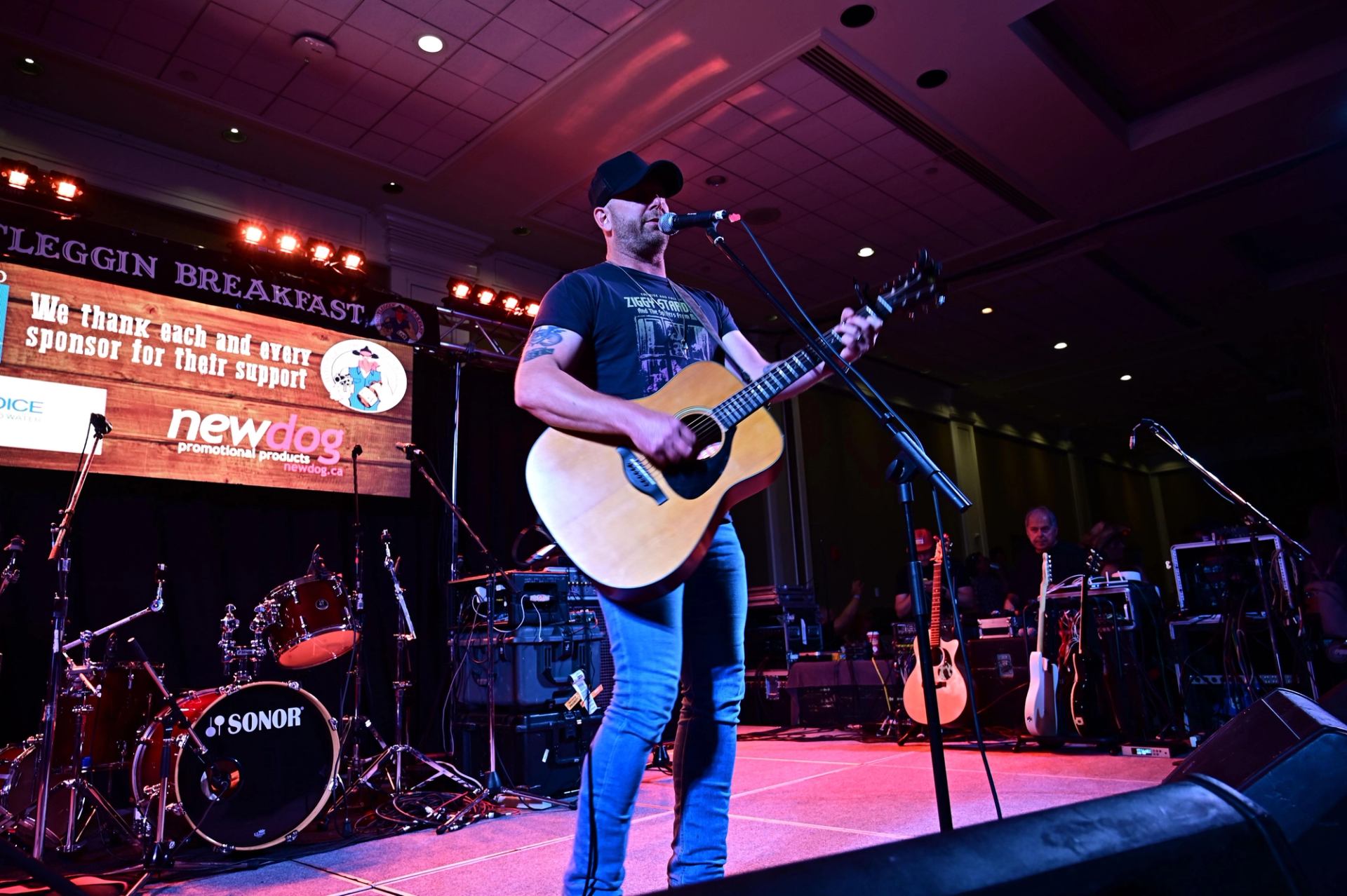 Guitarist sings into microphone on stage with drums, sponsor banner and stage lights behind..