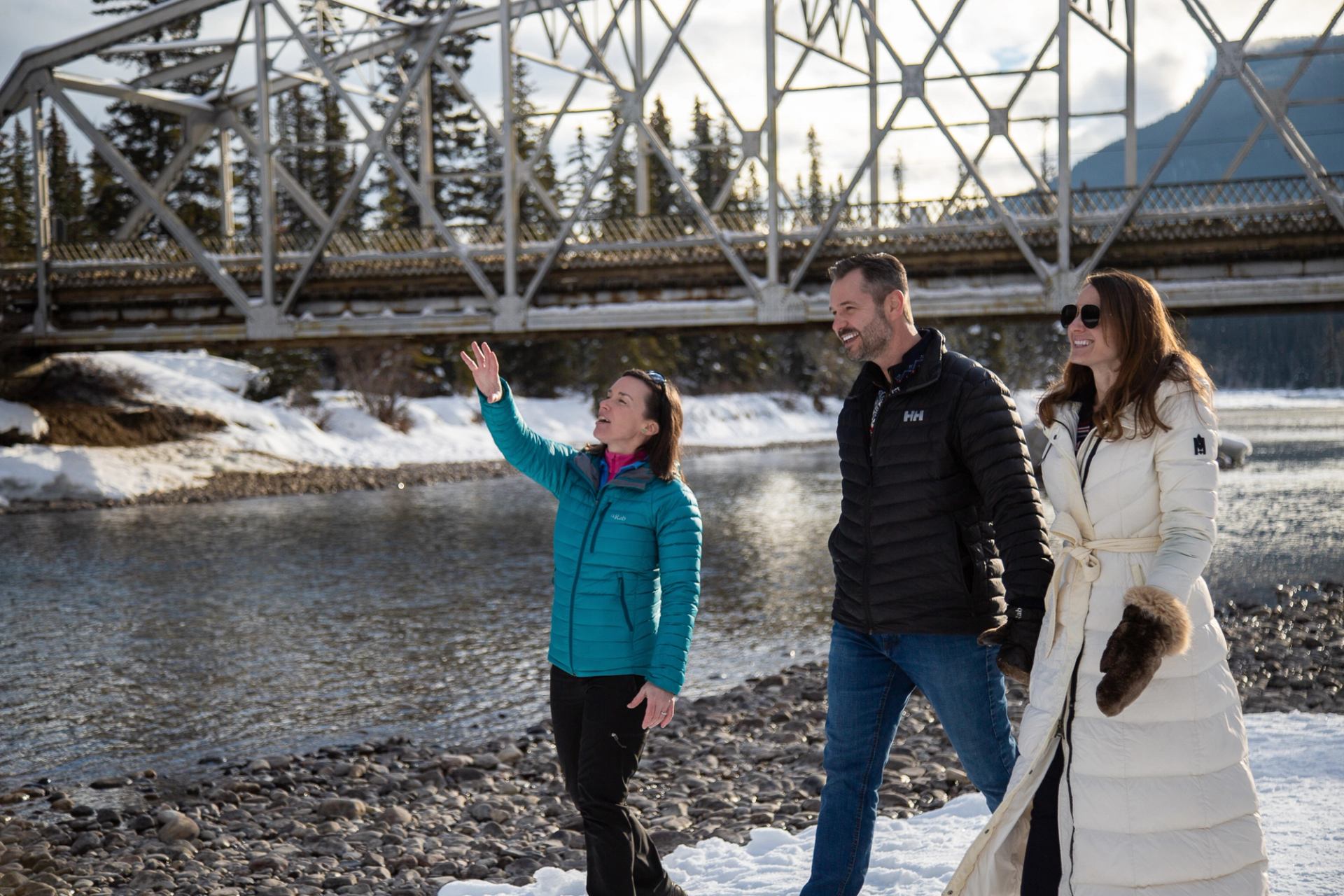 Three people walking on a snowy riverside path near a steel bridge with pine trees around