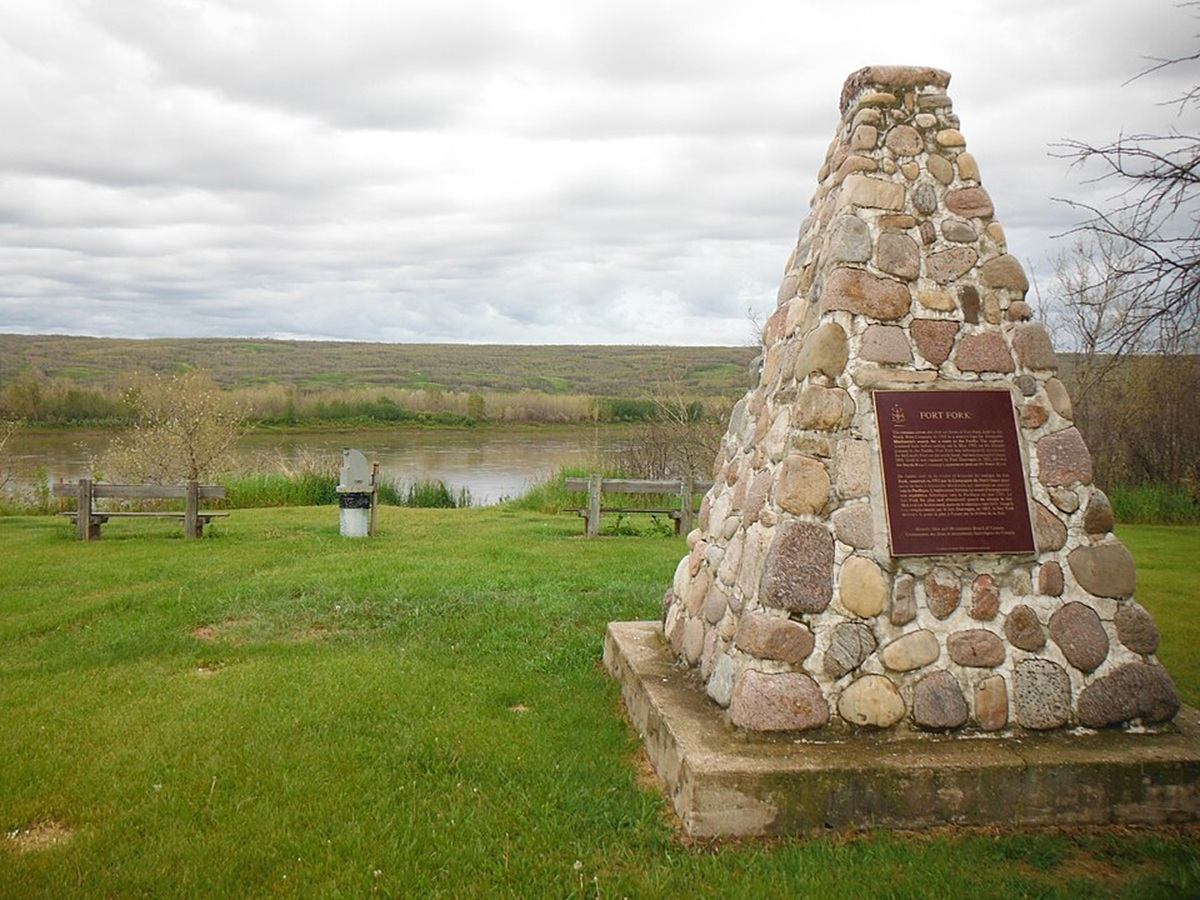Stone cairn monument with Fort Fork plaque beside river, benches and grassy field.