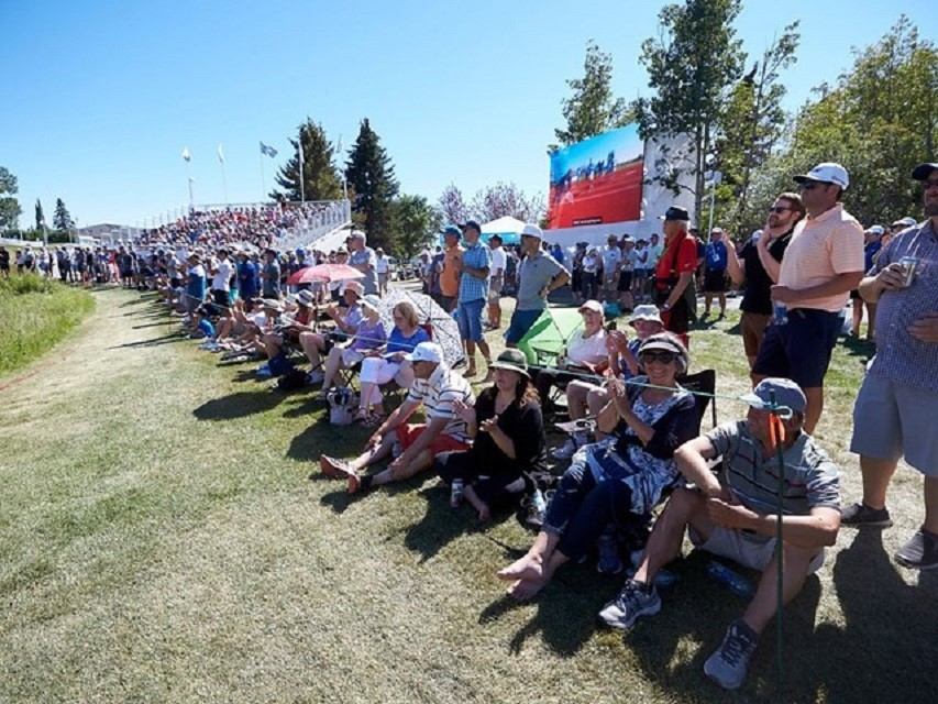 Spectators sit and stand on grassy slope beside bleachers at outdoor sporting event.