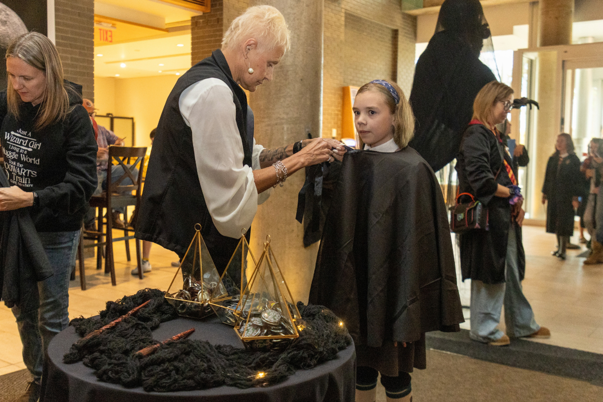 Costumed stylist fastens a black cape around a child beside a decorated table.