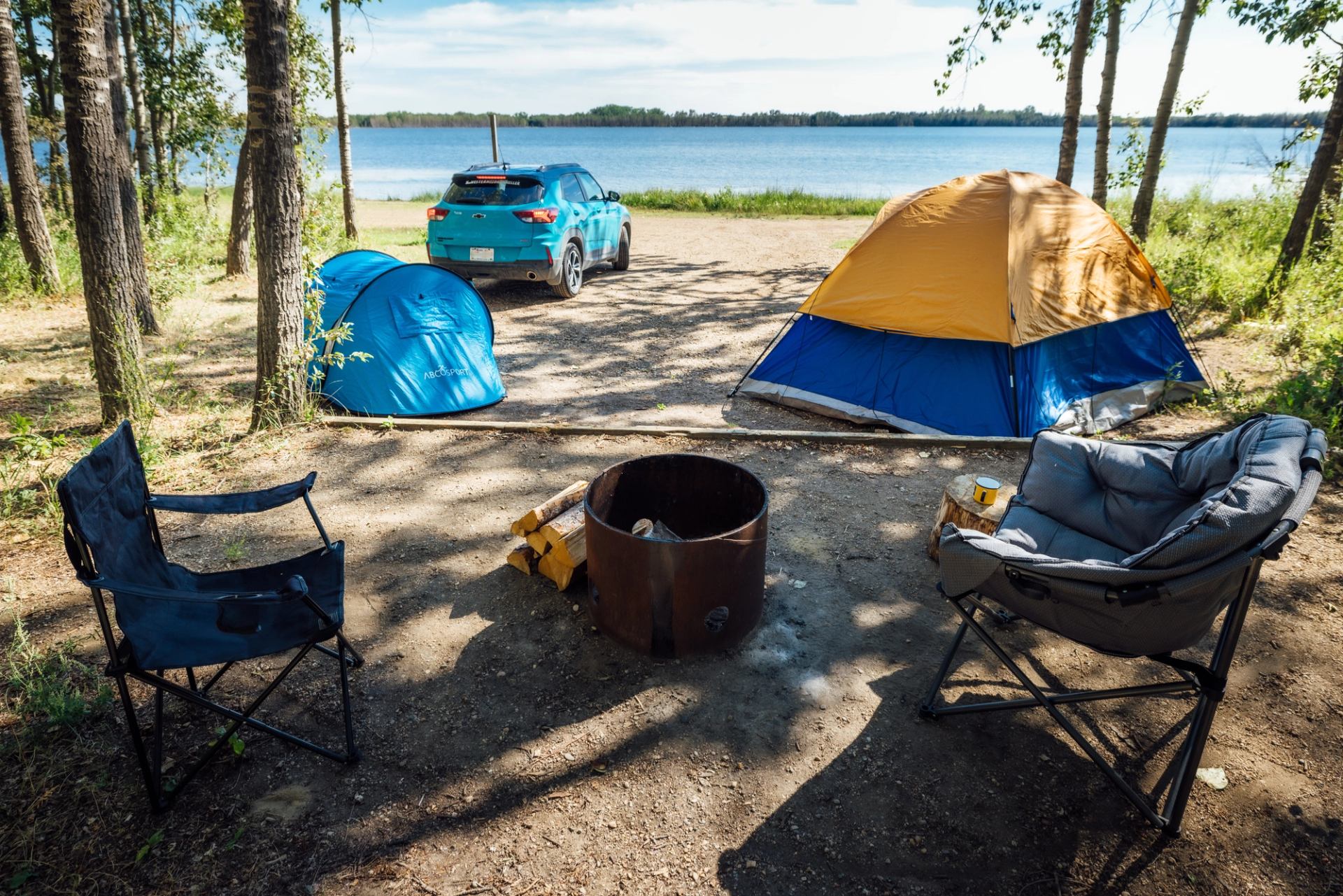 Camping site by a lake with a blue car, tents, chairs, and a fire pit with firewood.