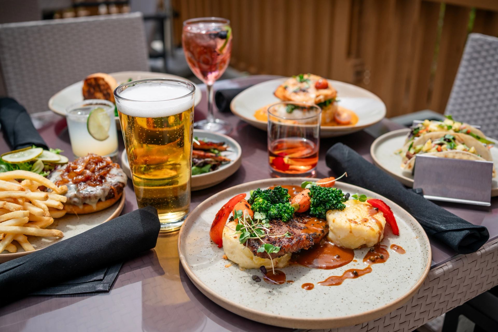 Steak with mashed potato dish, surrounded by other plates, and crisp drinks on the patio.