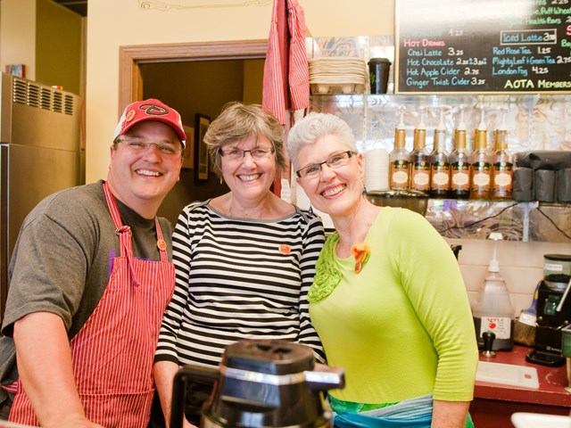 Three smiling people in a café with coffee gear and a menu board behind them.