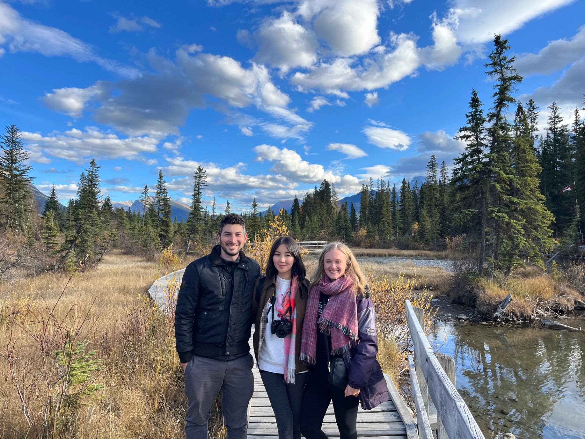 Three people stand on a wooden boardwalk in a marsh with mountains and trees.