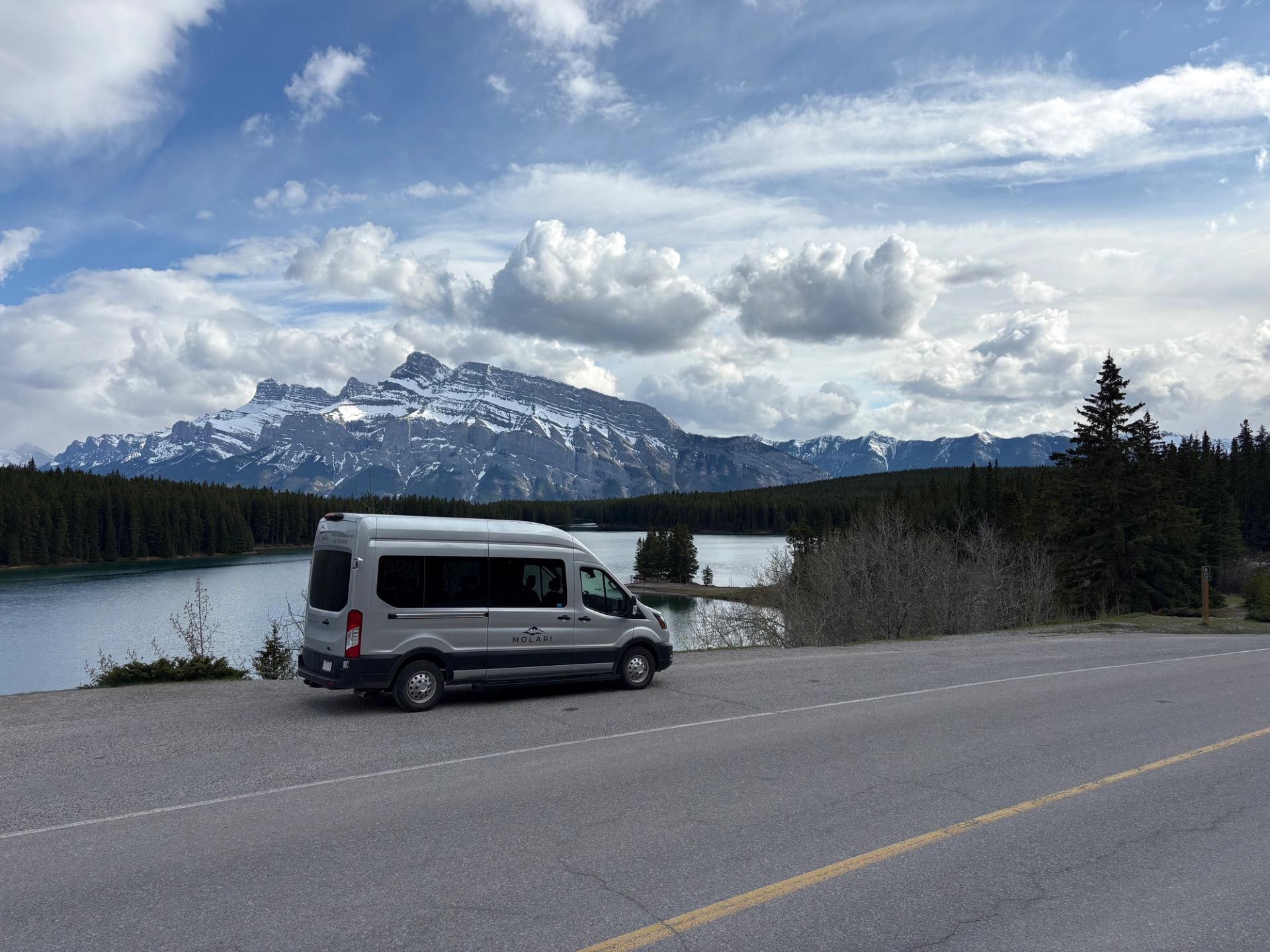 Silver passenger van parked beside lake with snow-capped mountains and evergreen forest.