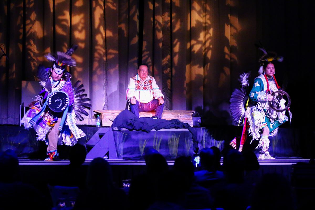 Stage performance with traditional attire, feathered headdresses, and tree-pattern backdrop.