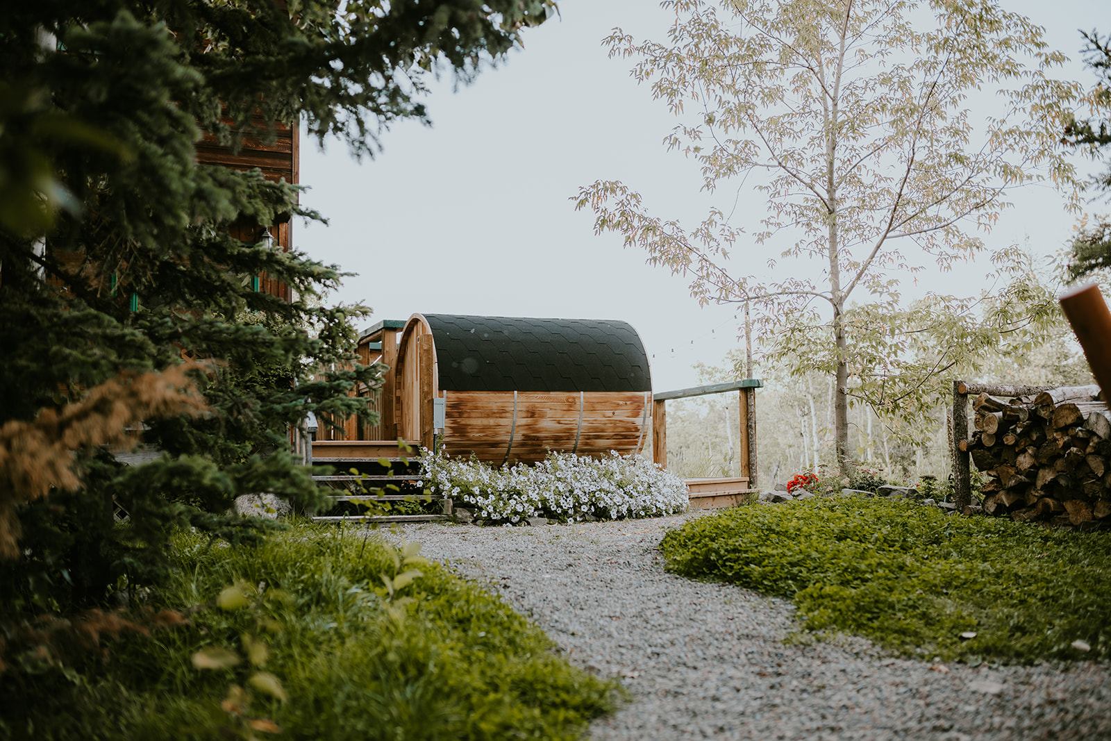 Barrel-shaped wooden sauna beside a gravel path, flowers, trees and stacked firewood.