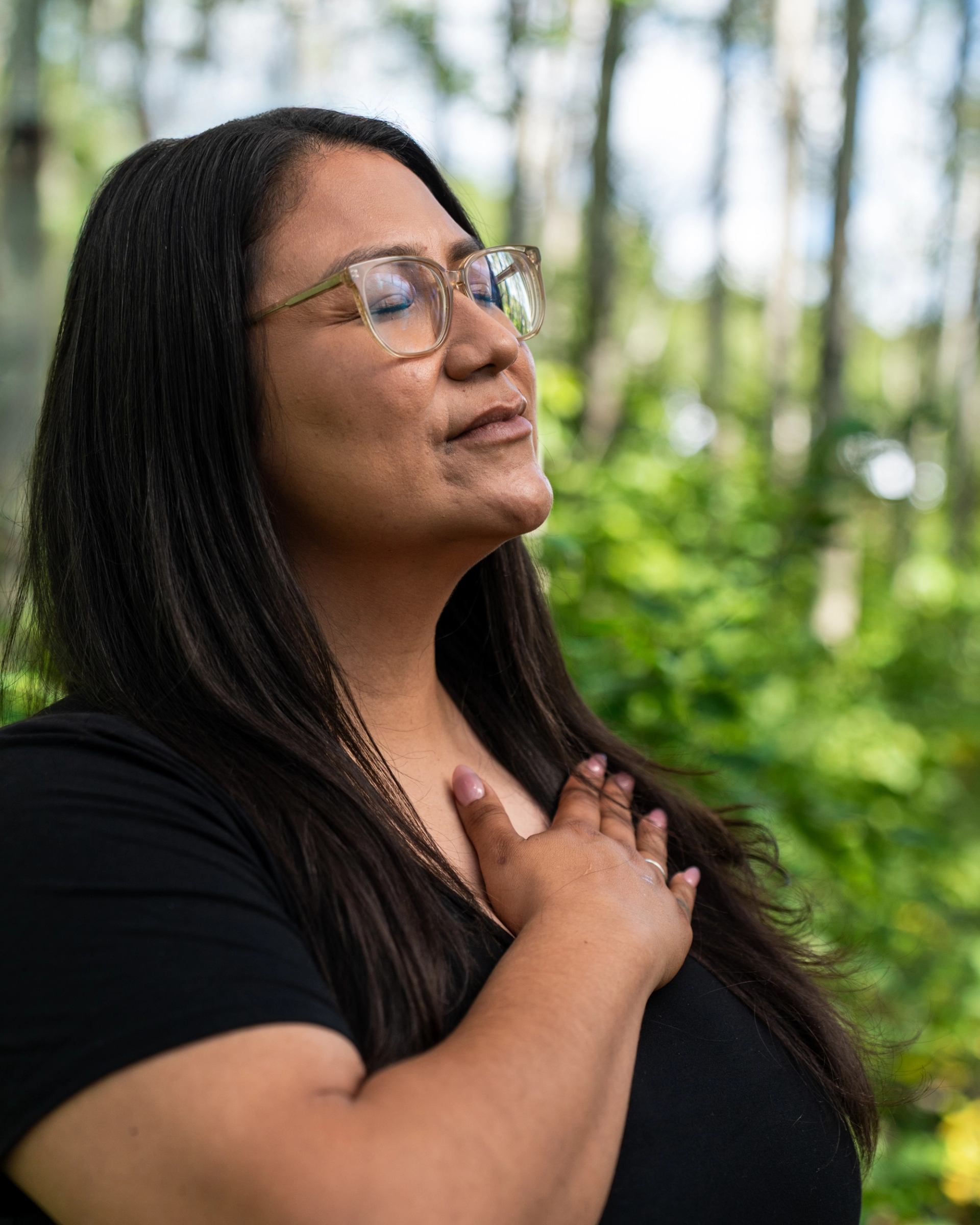 Person wearing glasses rests hand on chest with eyes closed in forest.