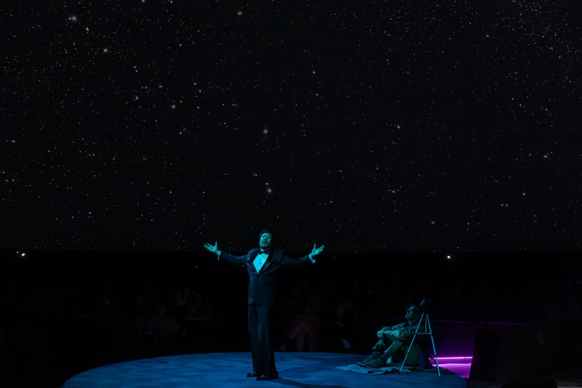 Performer on circular stage under starry sky with dramatic purple lighting.