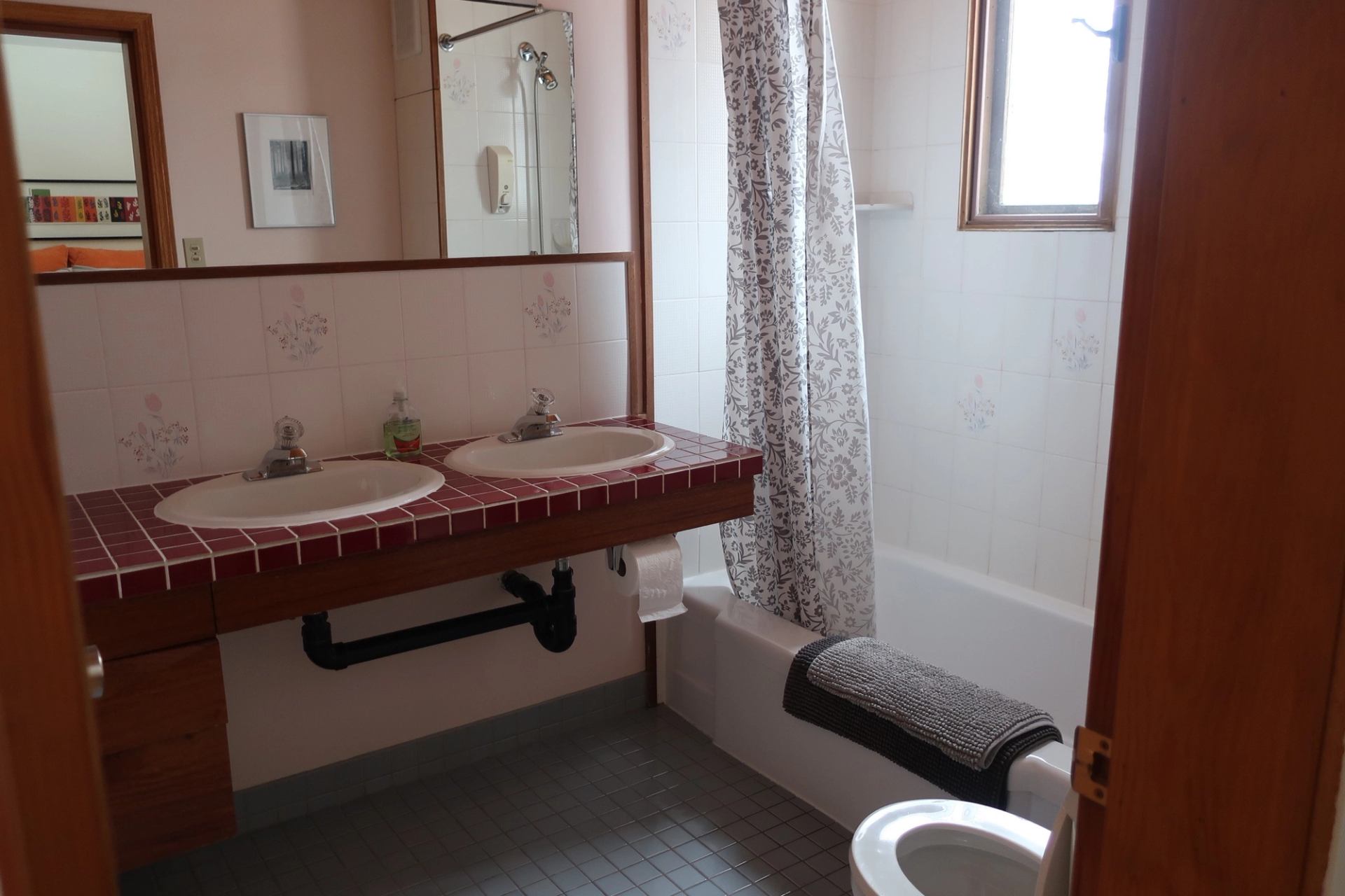 Bright bathroom with double sinks, red tile counter, and white-tiled tub with curtain.