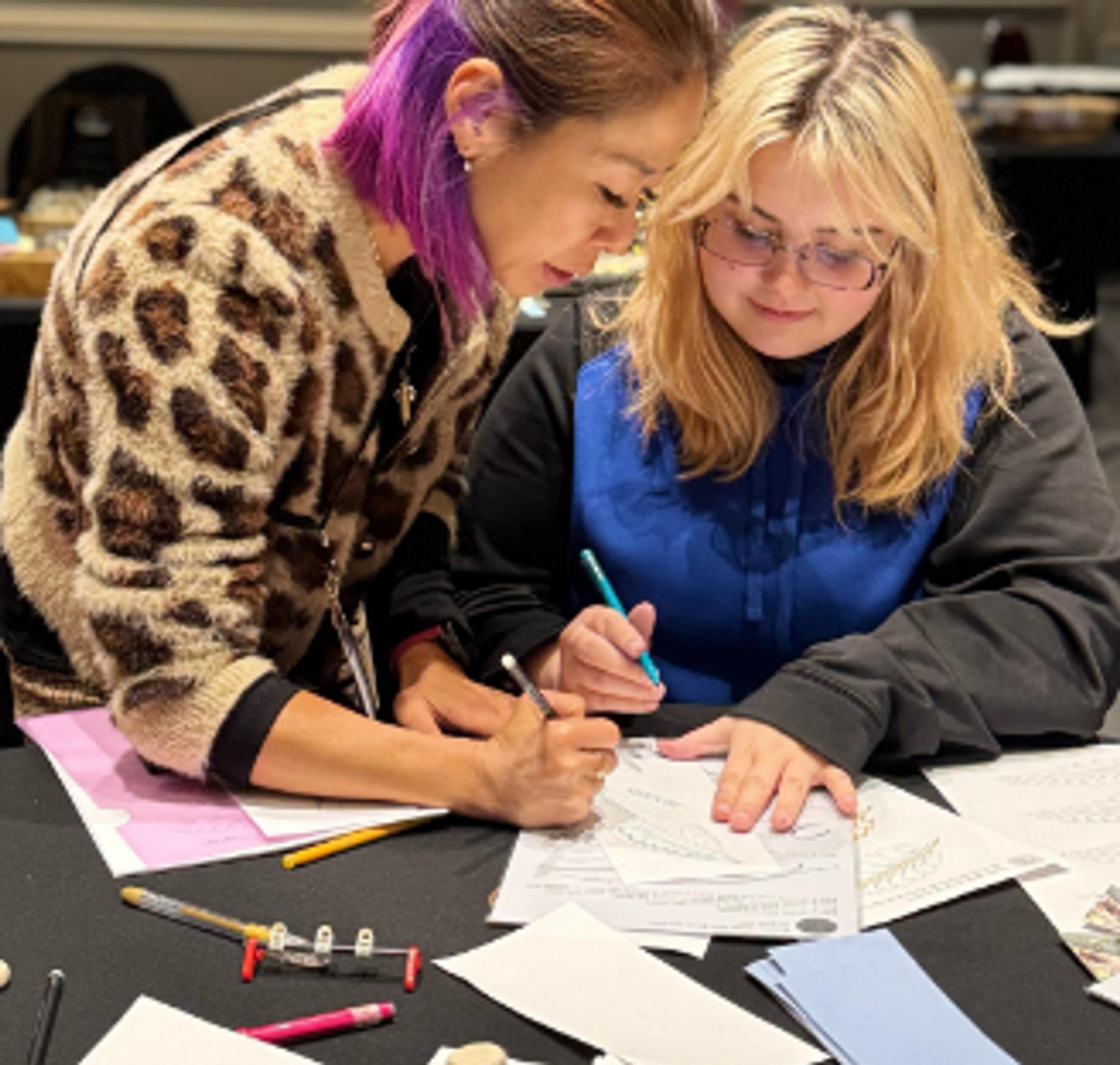 Two people write on papers at a table covered with art supplies.