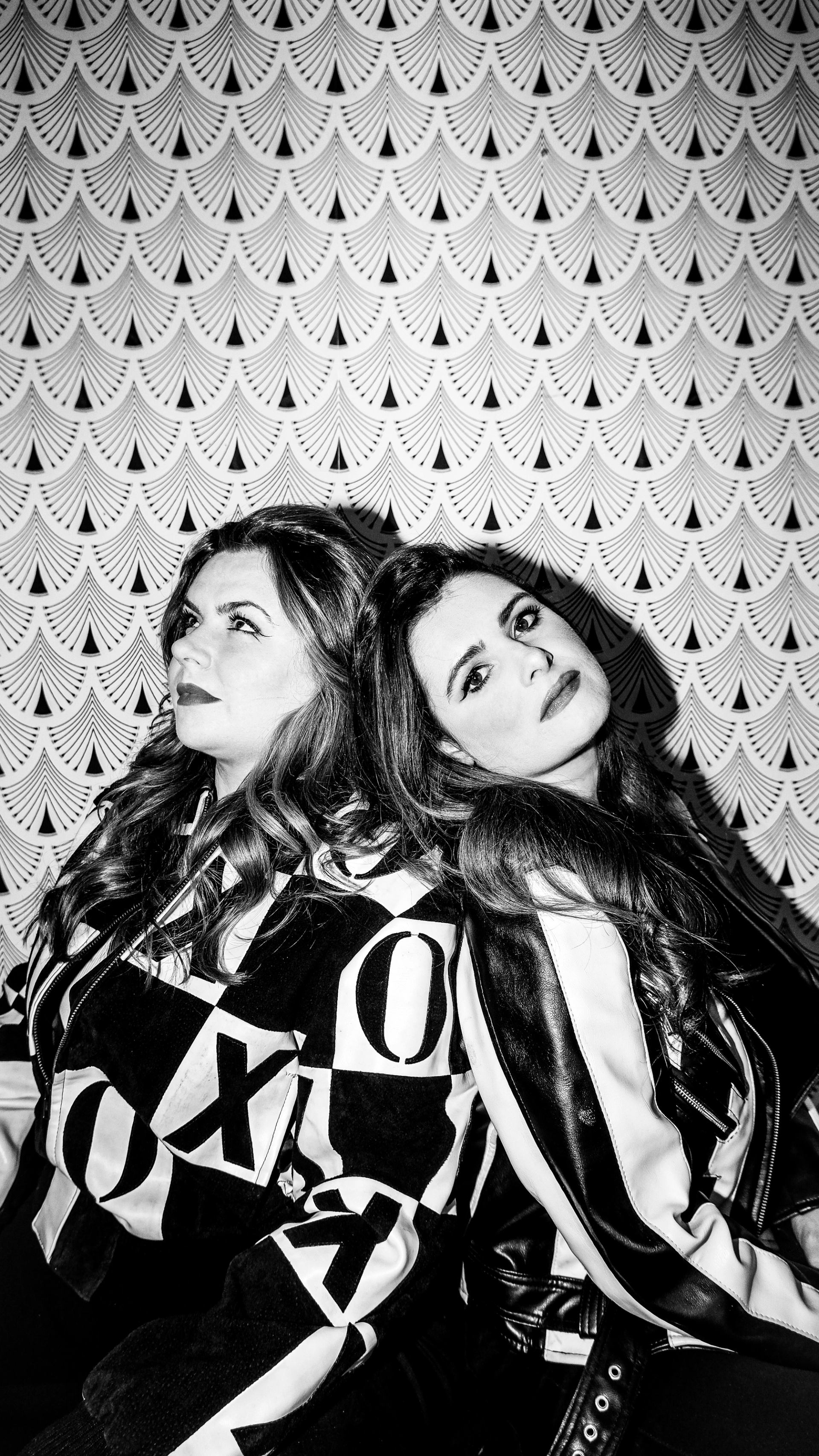 Two people sit back to back before patterned wall, wearing bold graphic jackets.
