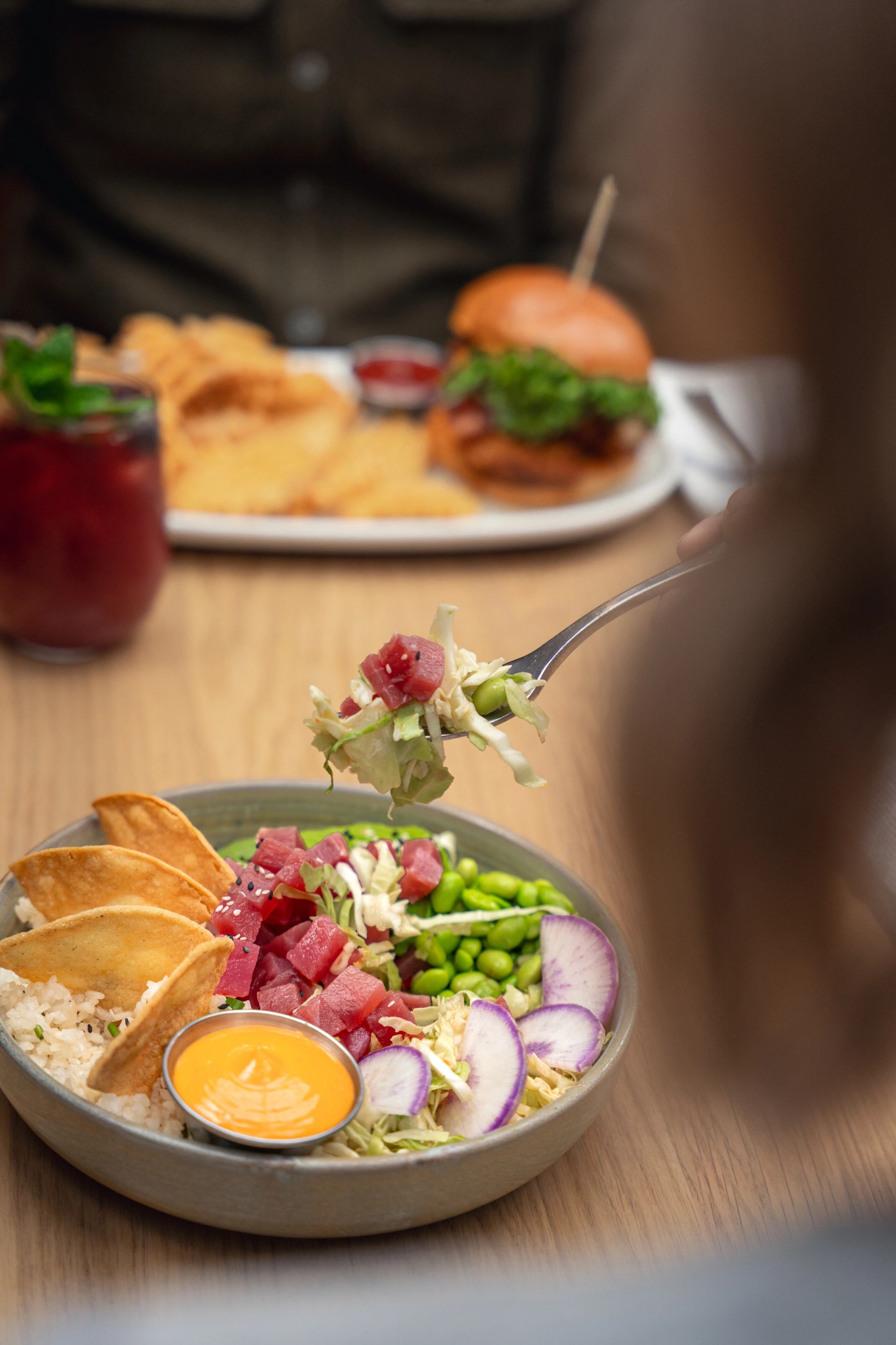 A person lifts a forkful of tuna and vegetables from a vibrant poke bowl with rice, edamame, radishes, and crispy chips.