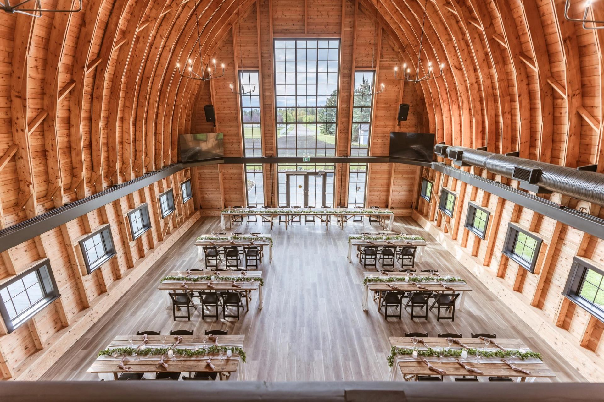 Spacious rustic event hall with wooden beams, large windows, and tables arranged for a gathering.