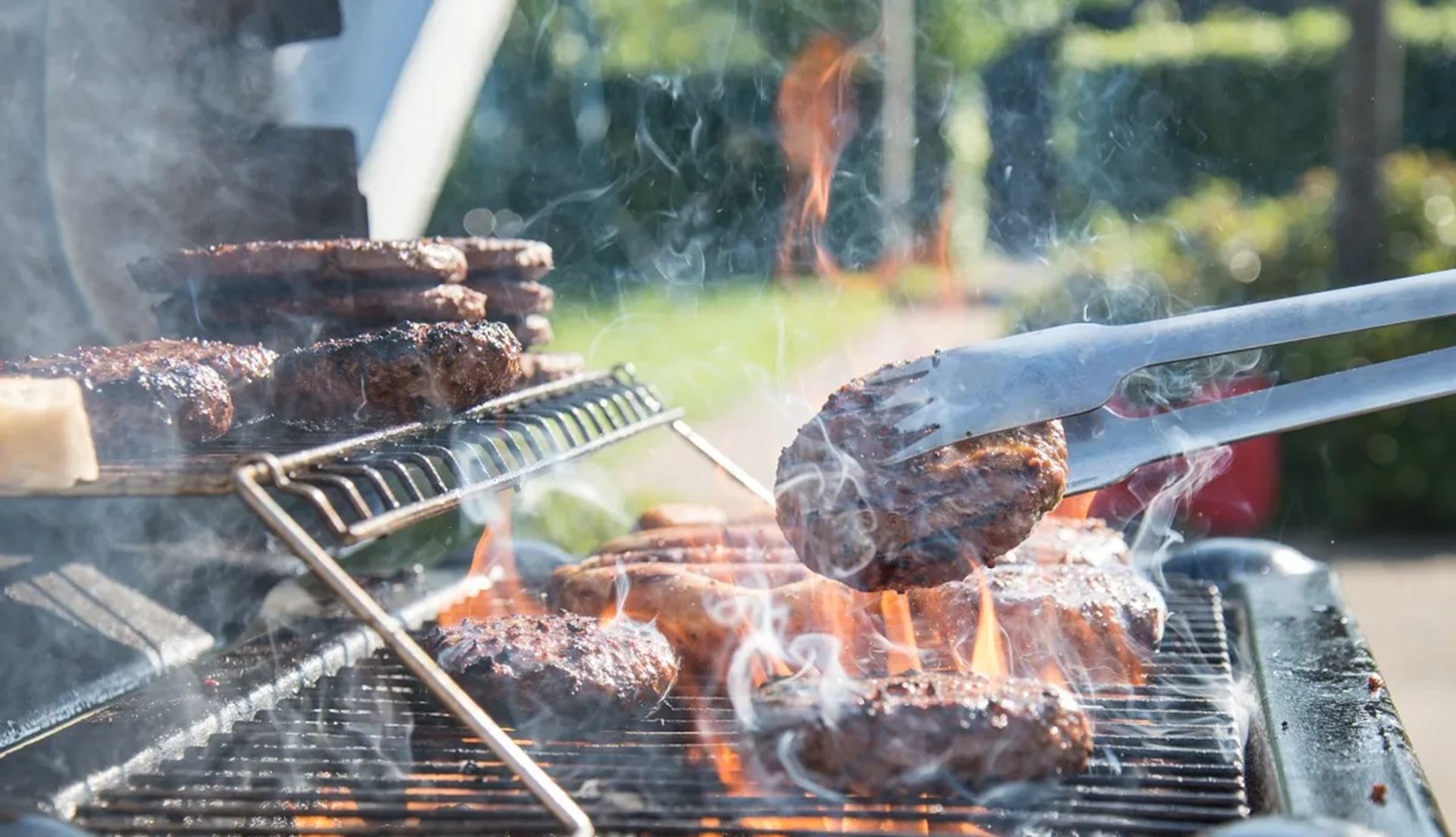 Hamburger patties cook on a flaming barbecue grill, with tongs lifting one patty.