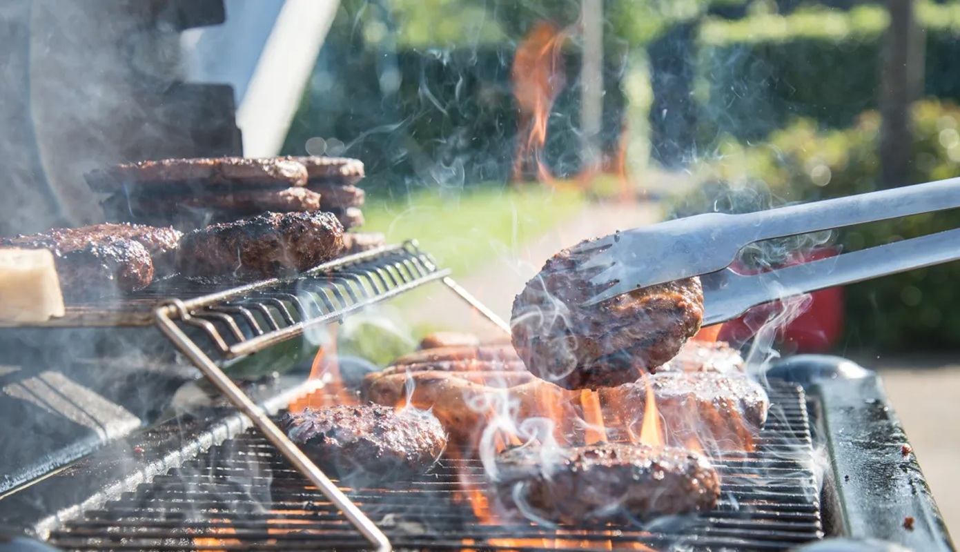 Hamburger patties cook on a flaming barbecue grill, with tongs lifting one patty.