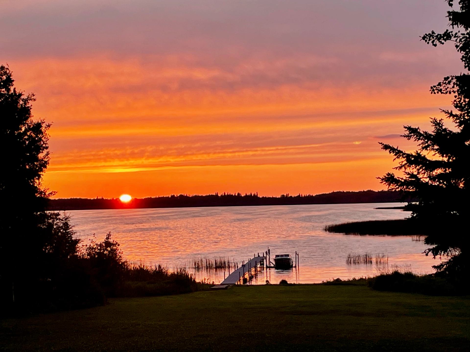Sunset over calm lake with docked boat, silhouetted trees and orange sky.