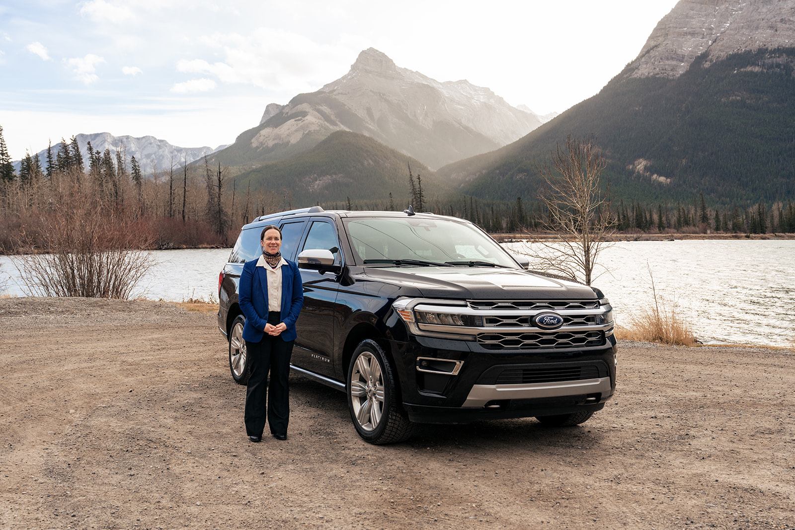 Individual standing beside a black SUV parked near a lake with tall mountains behind