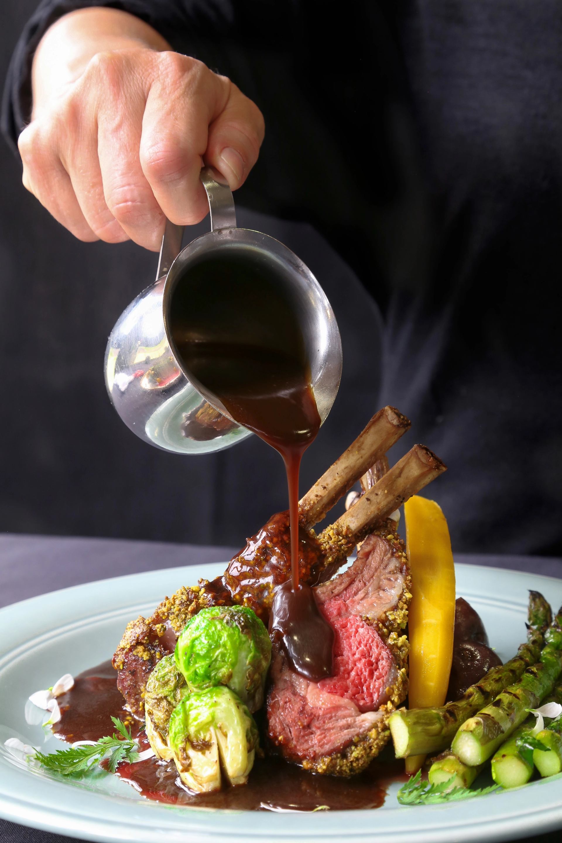 Rack of lamb topped with dark sauce, served with Brussels sprouts, asparagus, and a yellow vegetable slice.