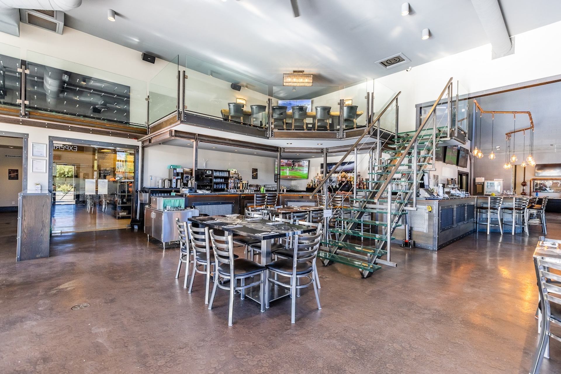 Spacious, multi-level restaurant with tables, a bar, and a mezzanine accessible by a metal staircase.