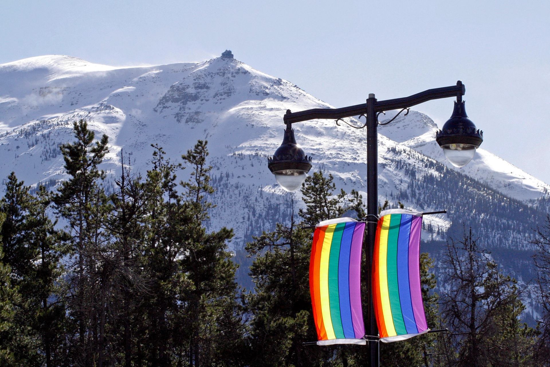 A rainbow banner hanging from a lamp post with snowy mountains in the background.
