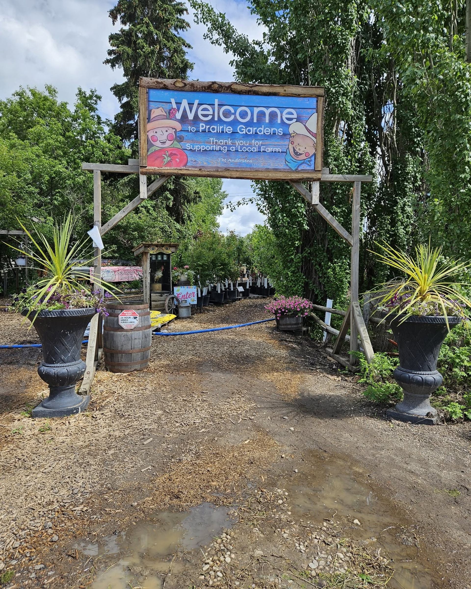 Farm entrance sign reading Welcome to Prairie Gardens above planters and muddy path.