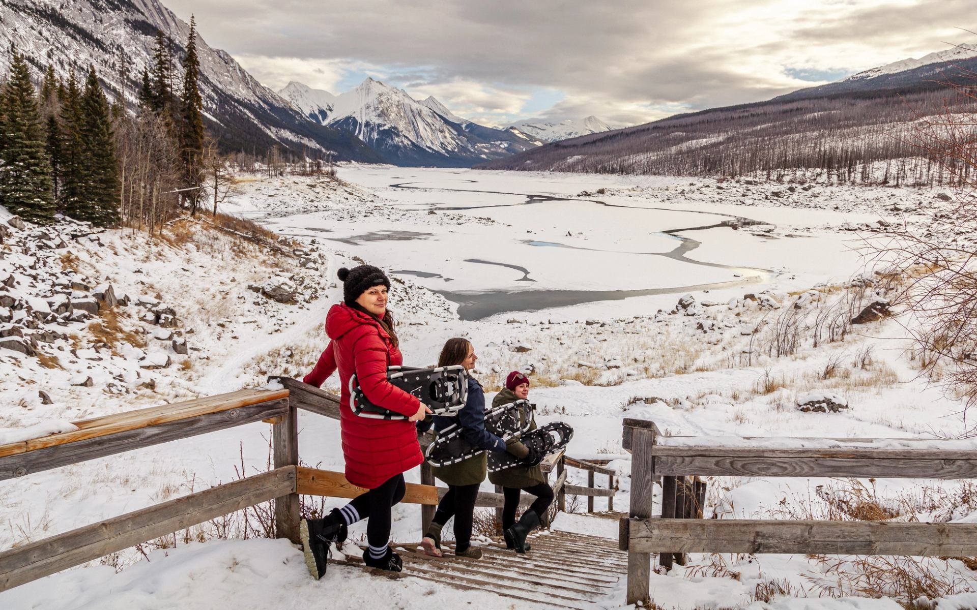 Group wearing snowshoes walking down wooden steps toward a frozen valley and snowy mountain range.