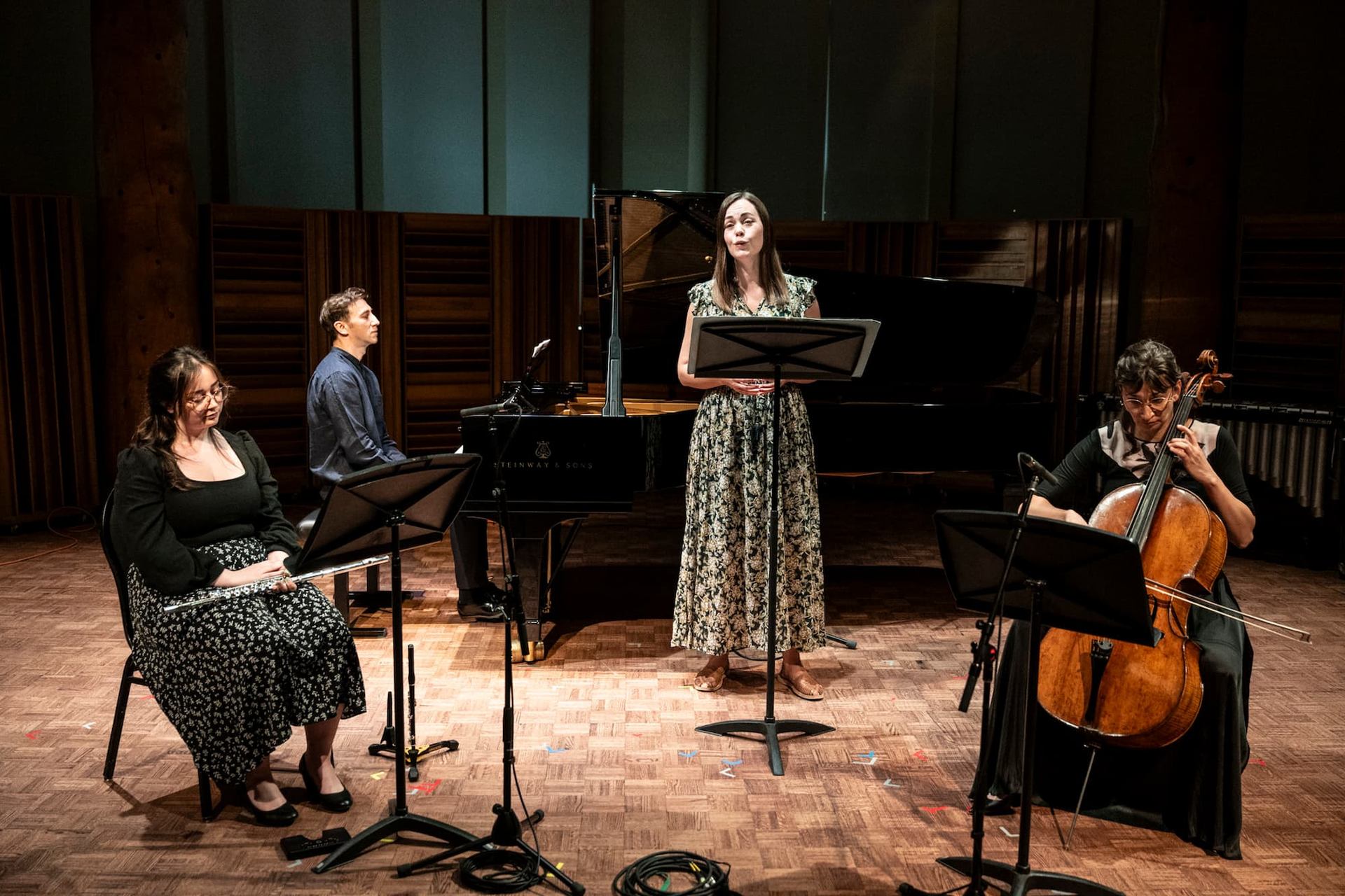 Musicians perform with piano, cello, flute and vocalist on a wood-floored stage.