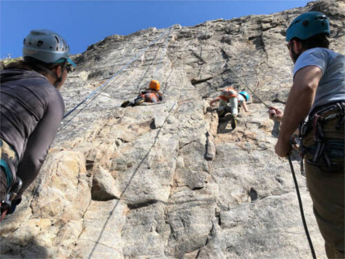 Rock climbers wearing helmets scale a steep cliff while belayers manage ropes below.