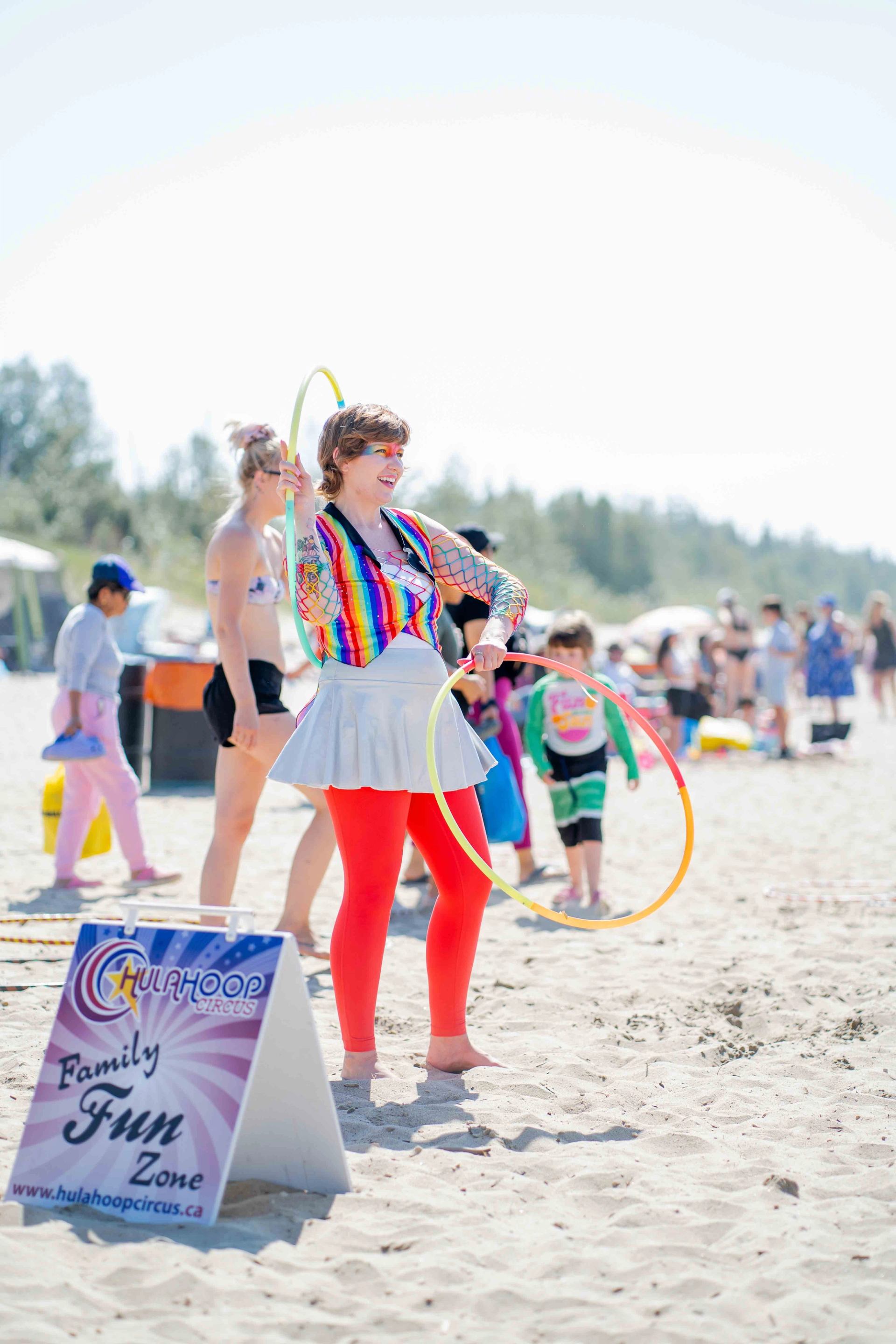 Person in colorful attire hula hoops on sunny beach near "Family Fun" sign, with families playing in festive atmosphere.