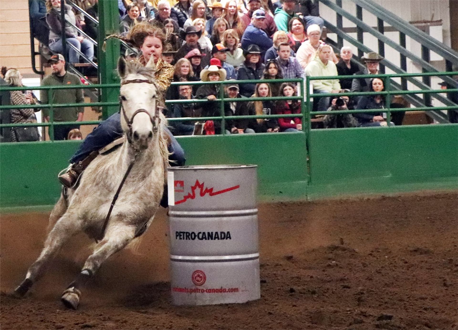 Rider on galloping horse rounding Petro-Canada barrel in indoor arena before spectators.