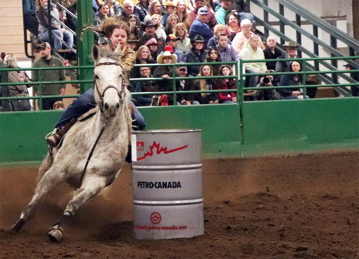 Rider on galloping horse rounding Petro-Canada barrel in indoor arena before spectators.