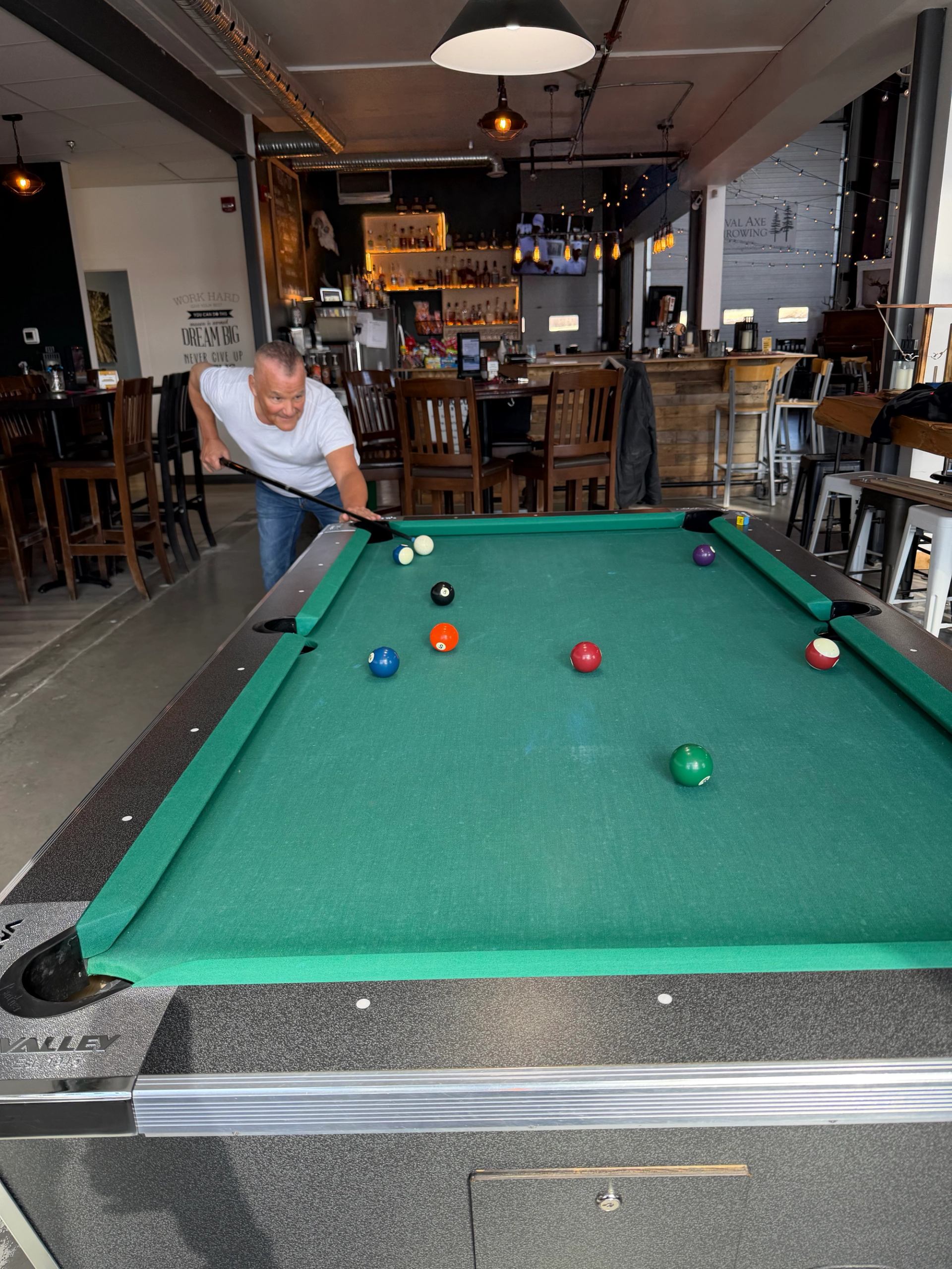 Man in white T-shirt taking pool shot on green table inside bar.