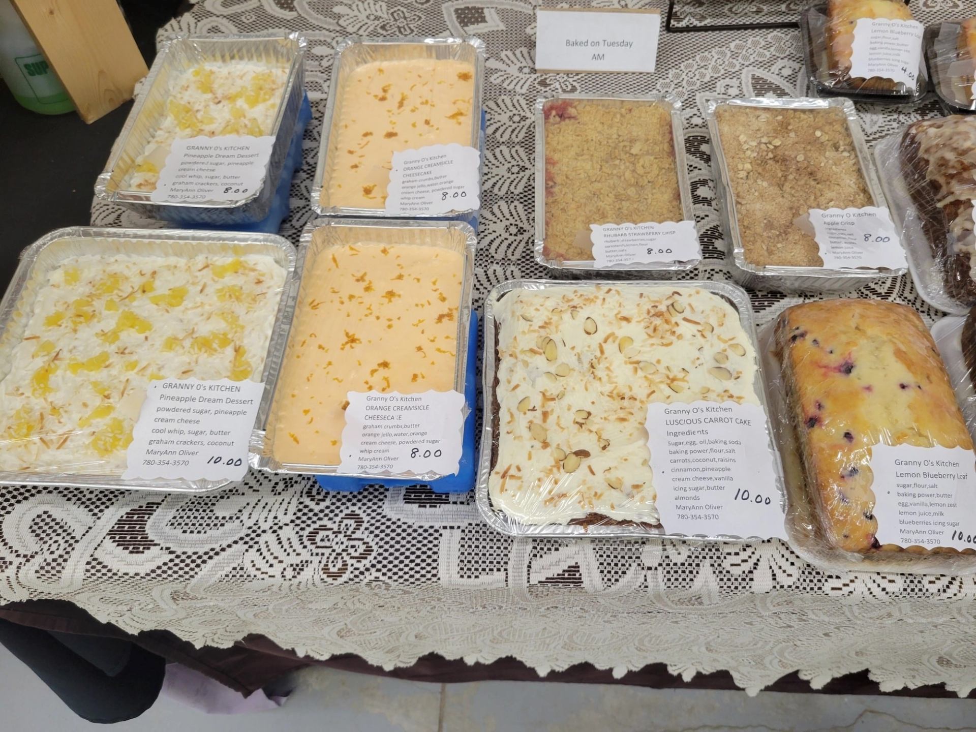 Display of homemade cakes and desserts in foil trays on a lace‑covered market table.
