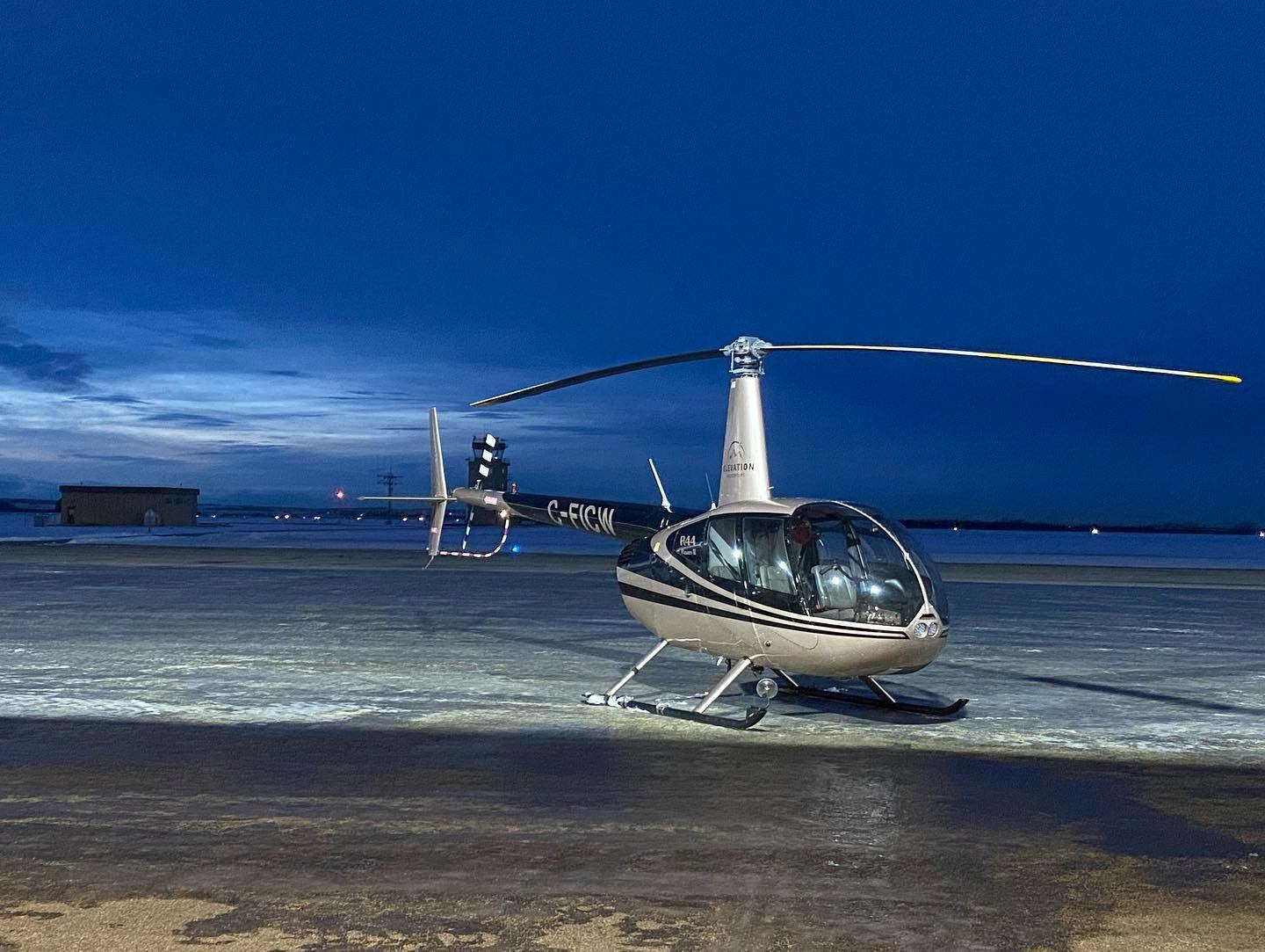 Silver helicopter on wet tarmac at dusk under a dark blue sky.