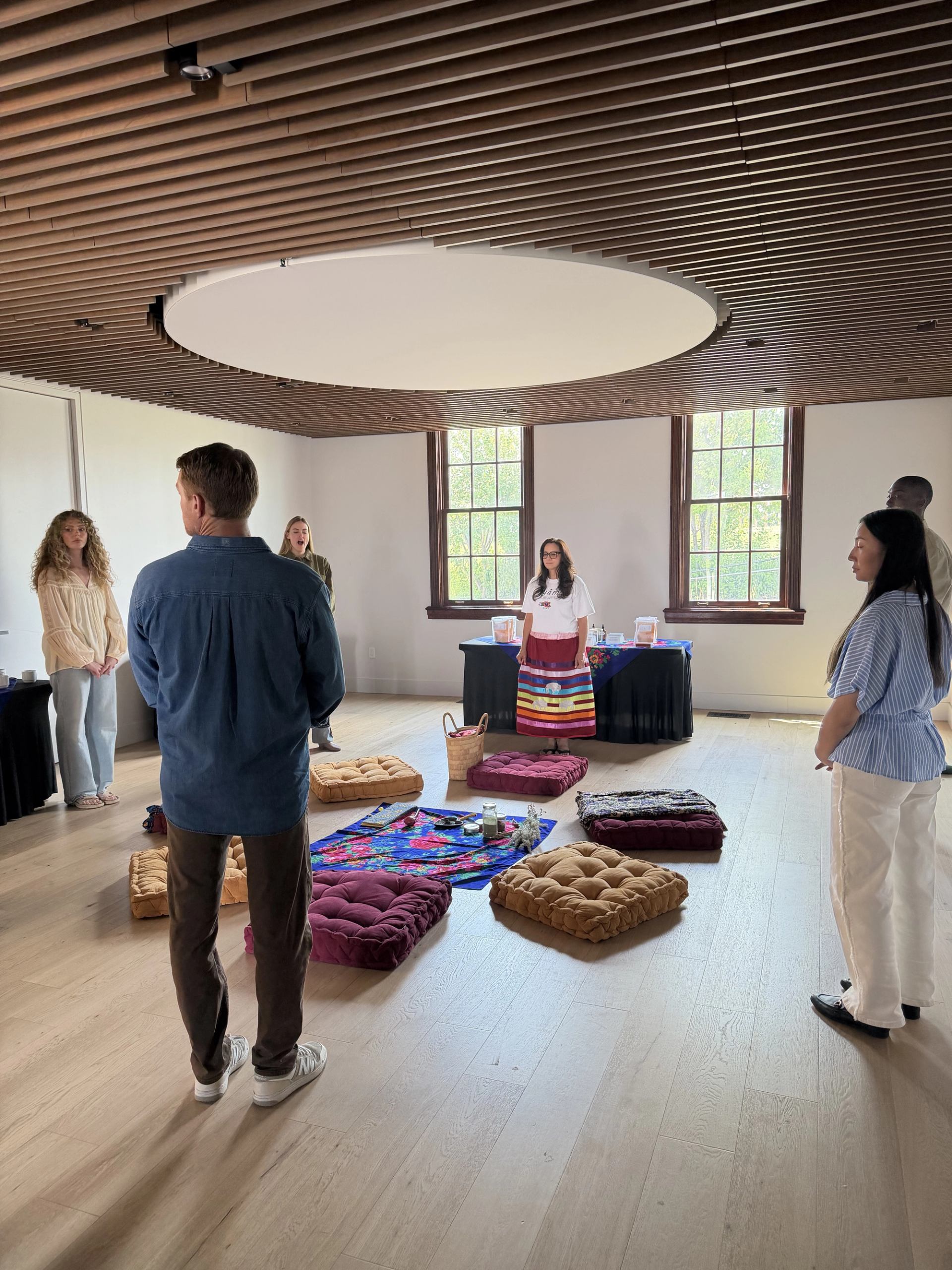 People stand in a circle around floor cushions and blankets in a bright room.
