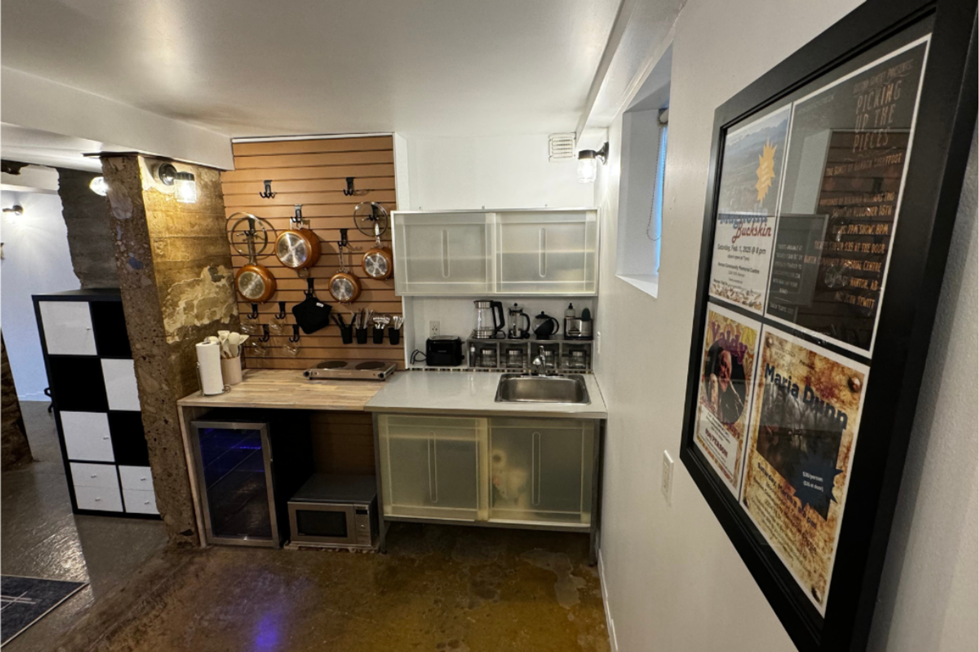 Compact kitchenette with hanging copper pots, small appliances, sink and framed posters on wall.
