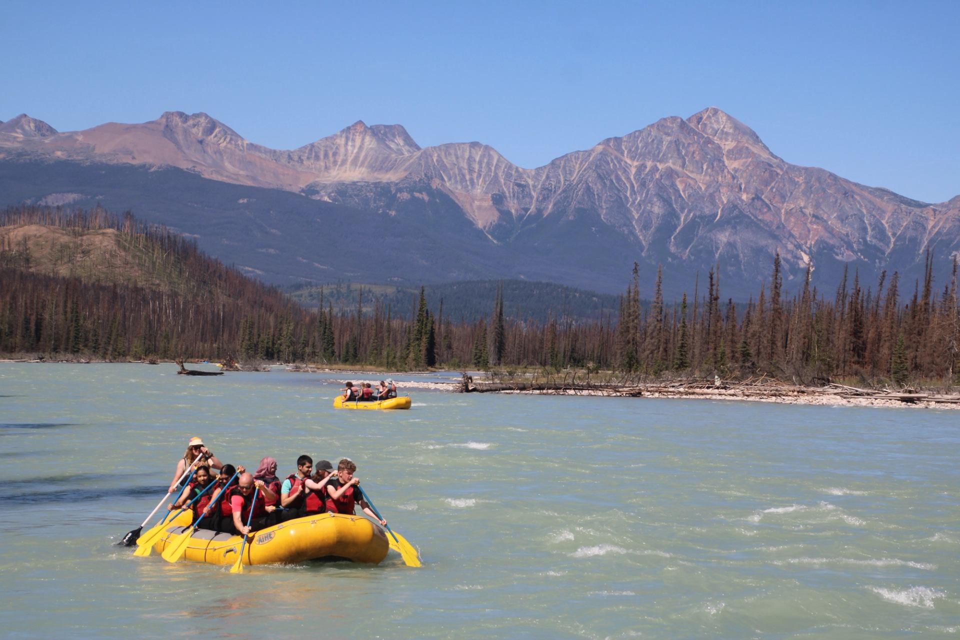 Yellow raft moving down a wide pale‑blue river with tall mountains and trees in the background.