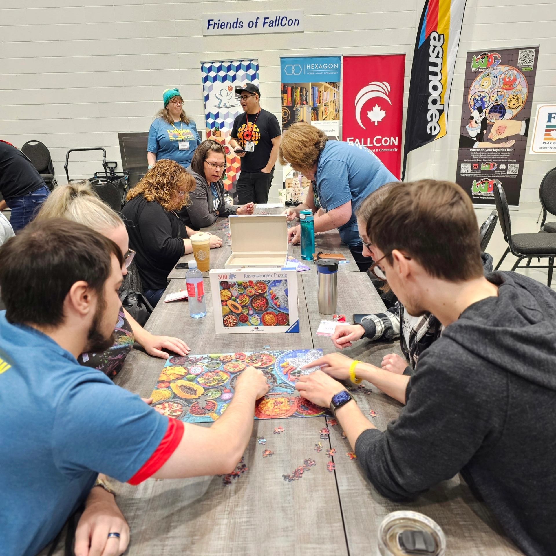 People at FallCon event assembling a colourful Ravensburger jigsaw puzzle at long table.