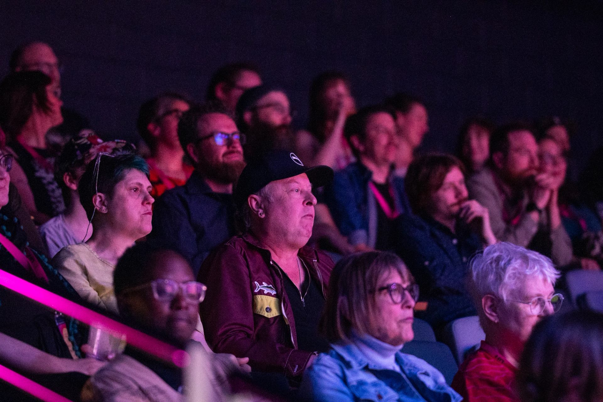 Audience members sit in dim theatre seating, watching an unseen event under purple lighting.