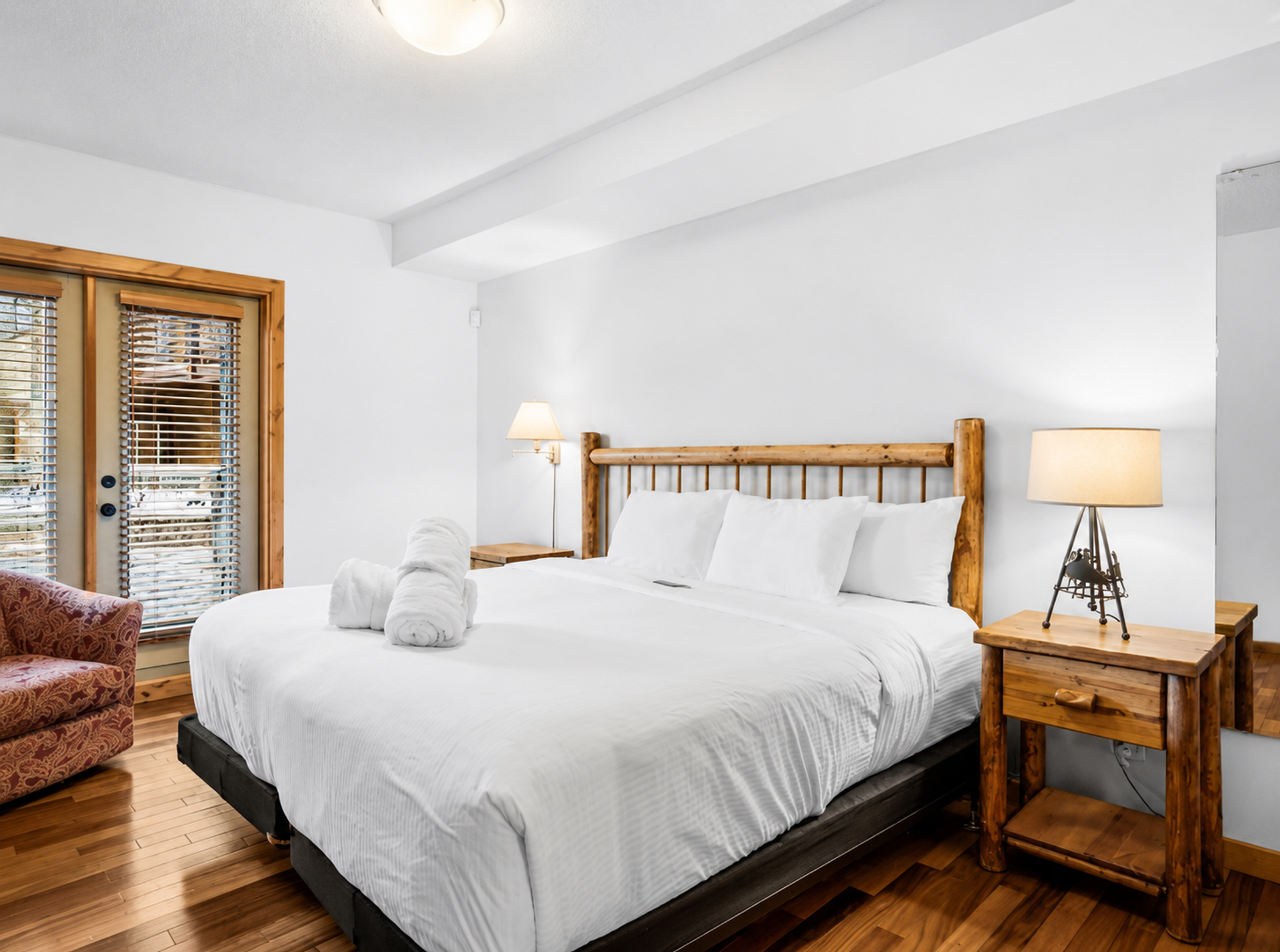 Bedroom with wooden bed frame, white bedding, bedside lamps and glass doors with blinds.