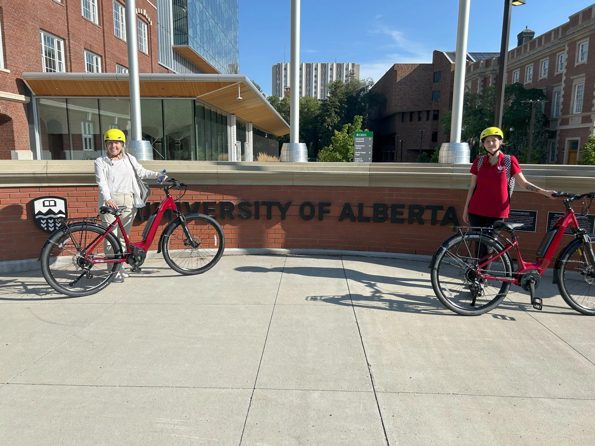 Two helmeted cyclists pose with bikes beside “University of Alberta” sign on sunny day.