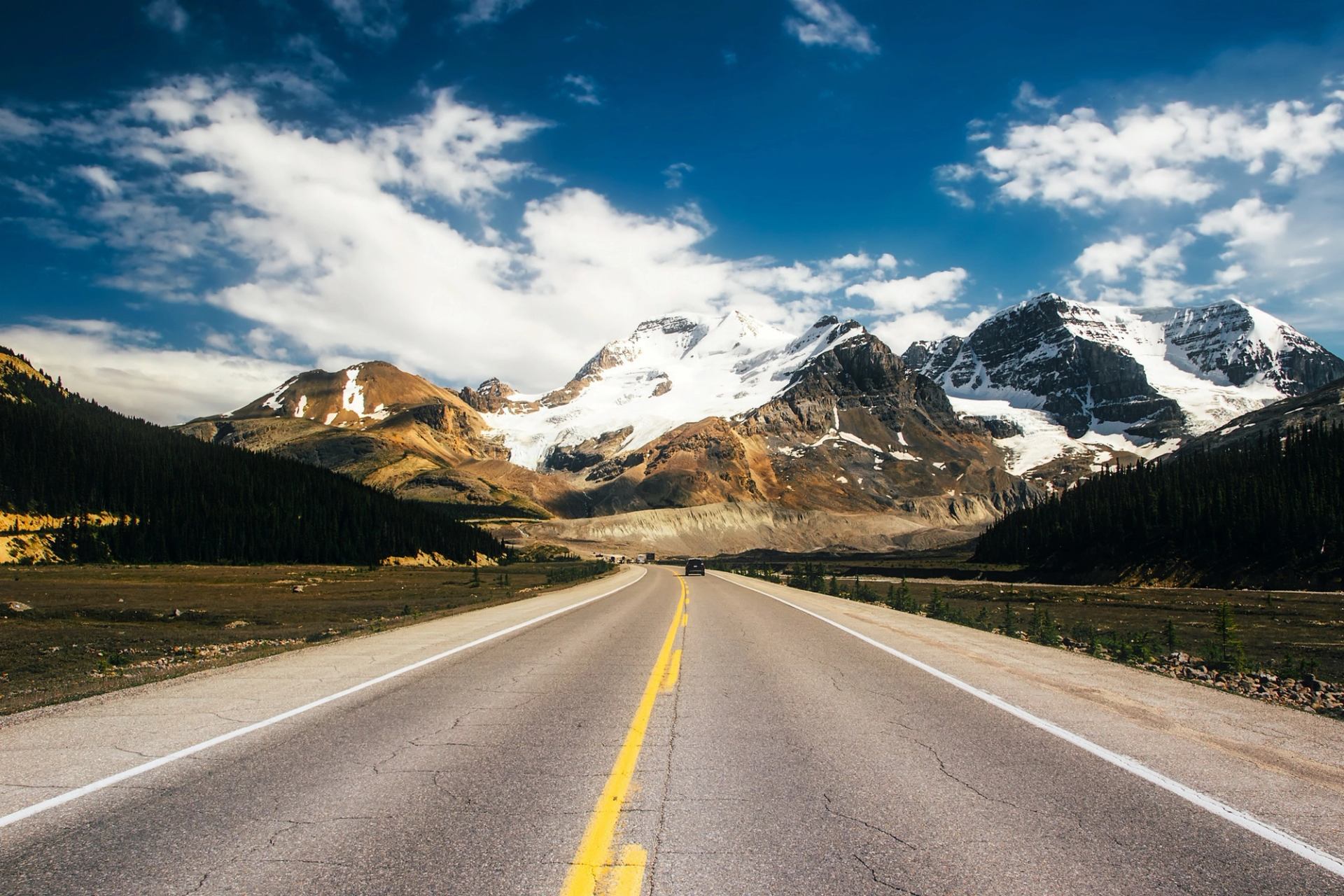 Highway leads toward snow-covered mountains beneath a blue, cloud-filled sky.